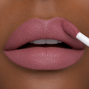 Close-up of a woman's lips with a medium-dark skin tone applying Runway Rogue Velvet Glam long lasting matte liquid lipstick in Glam Squad to her lips, a faded mauve color with a hint of plum